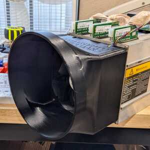 Black circular-mouth shroud mounted to silver equipment with exposed circuit boards and ribbon cables.